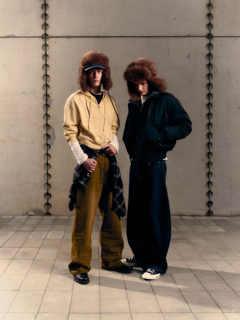 Two models in trapper hats and layered streetwear—hoodie, bomber and wide-leg jeans.