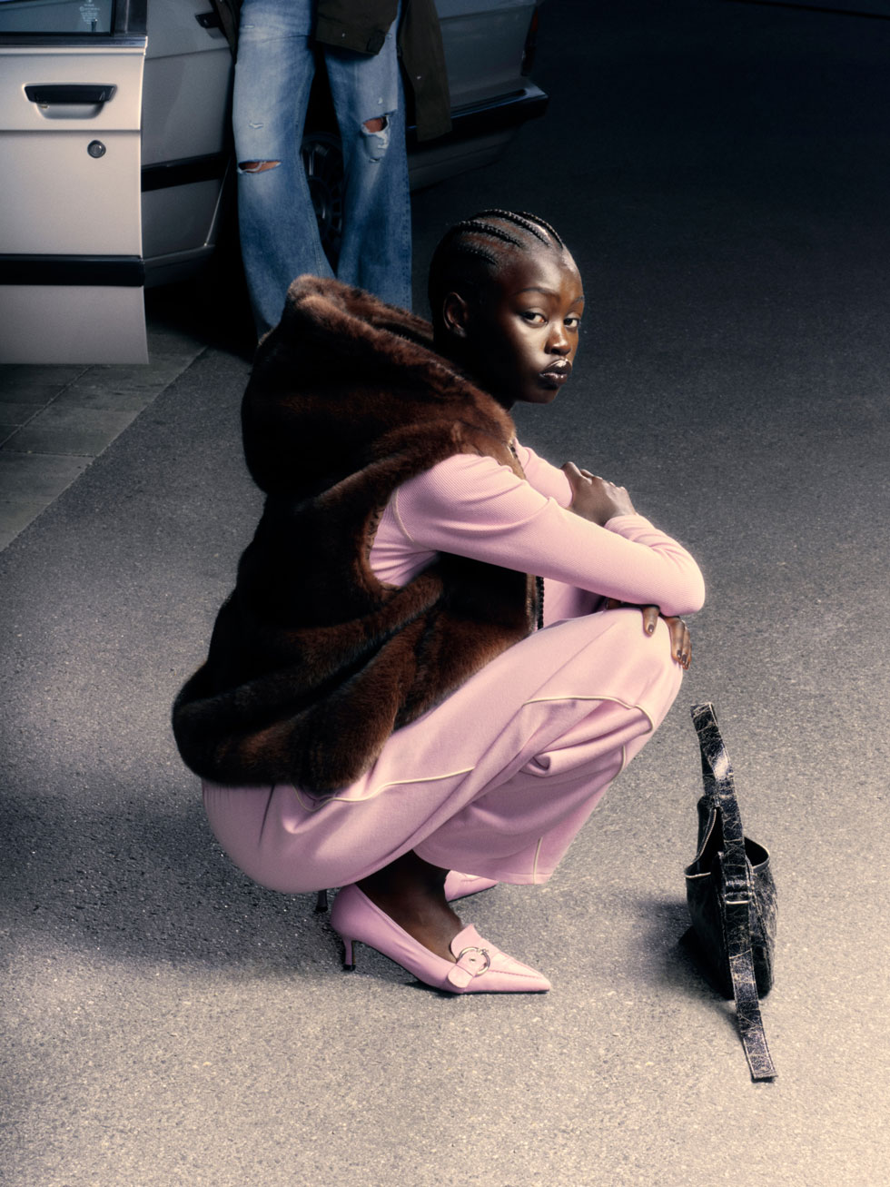 Model crouches on street in a pink set with a brown faux-fur vest and pink kitten heels.