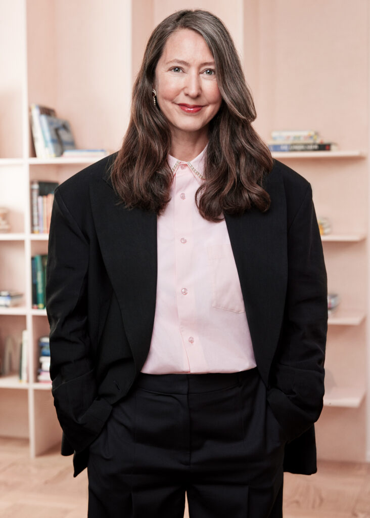 Headshot photo of Ann-Sofie Johansson wearing a black suit with a pink bookshelf in the background
