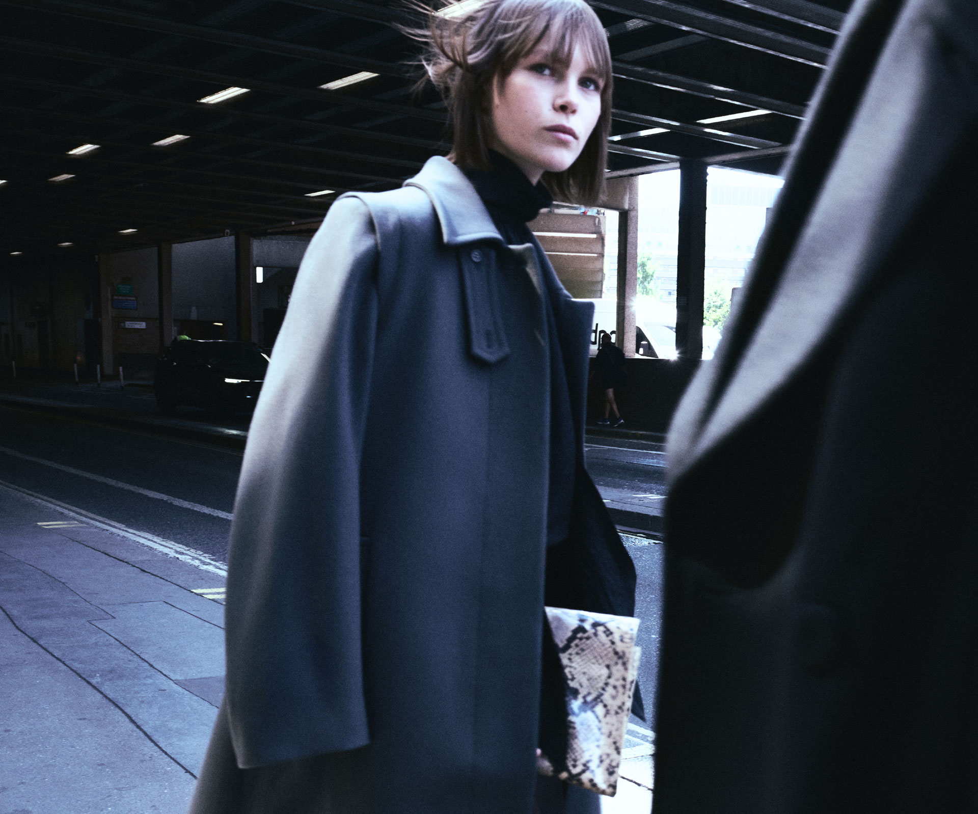 Model in a charcoal overcoat and black turtleneck holds a snake-print clutch while walking under an overpass.