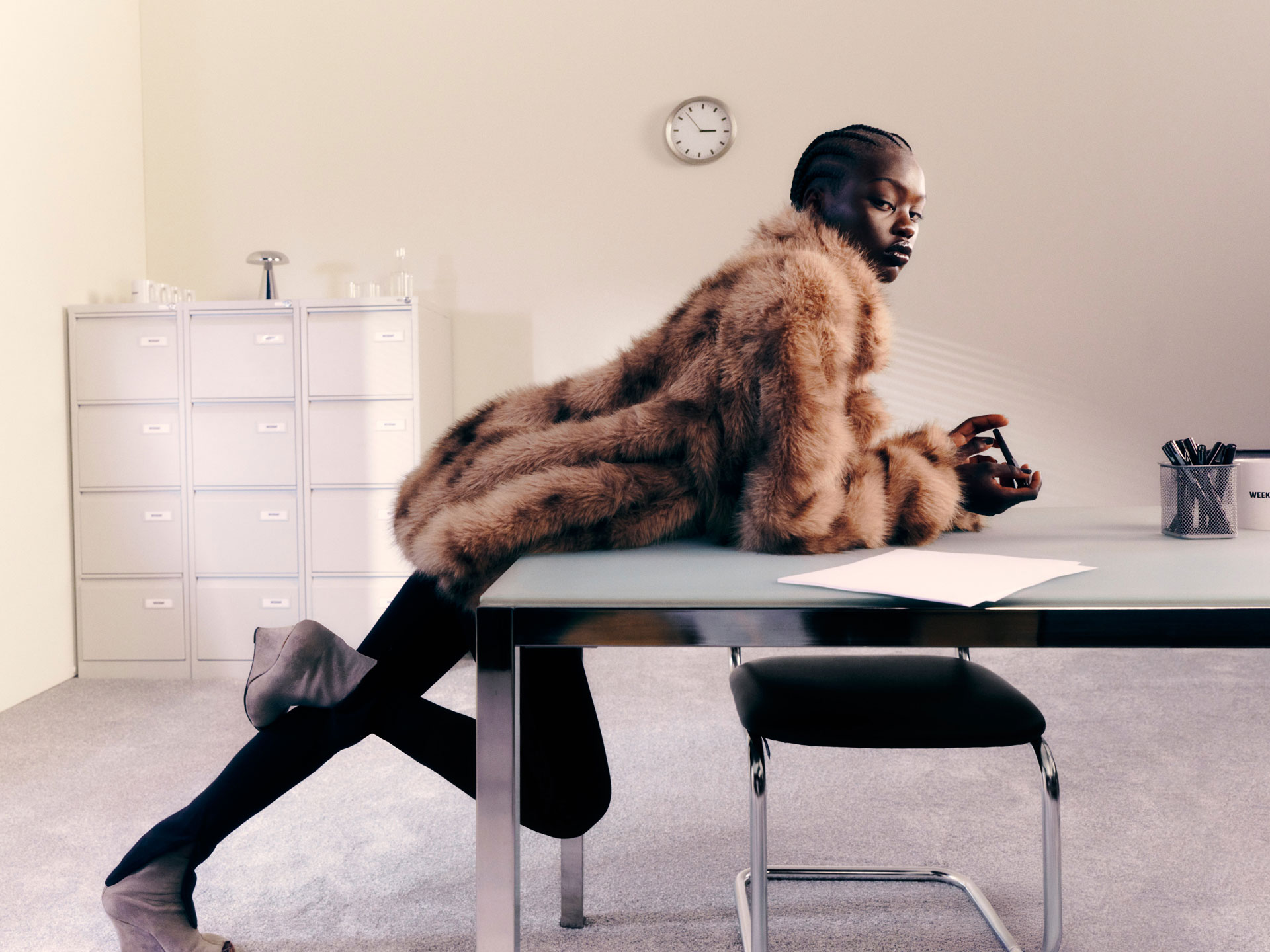 Model in tan faux-fur coat leans across a glass table; clock and filing cabinets behind.
