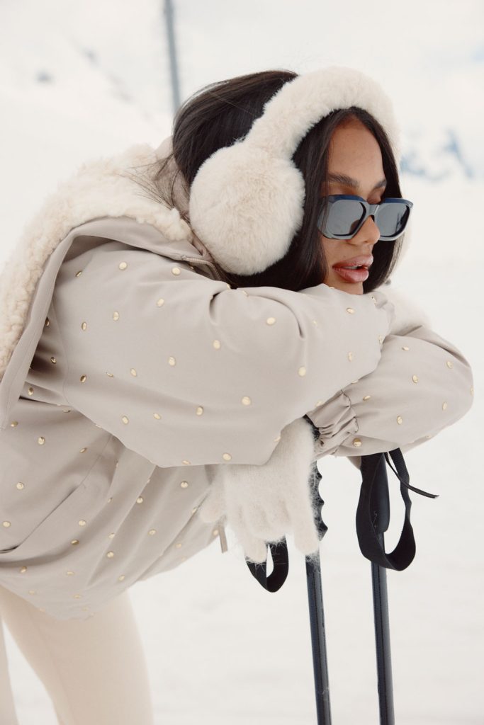 Model leaning on ski poles, wearing a beige studded jacket with faux shearling details, cream earmuffs, and black sunglasses against a mountain backdrop.