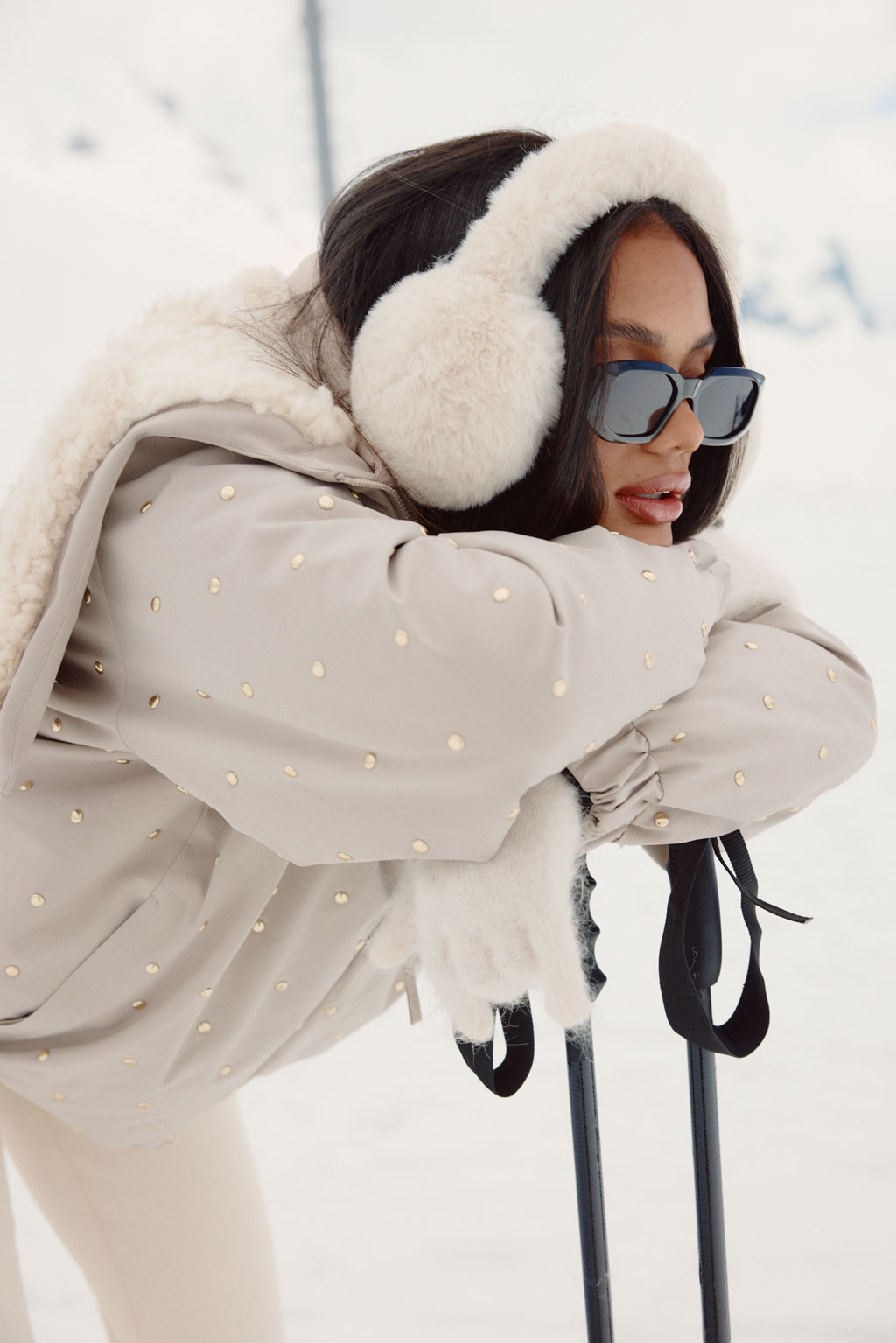 Model leaning on ski poles, wearing a beige studded jacket with faux shearling details, cream earmuffs, and black sunglasses against a mountain backdrop.