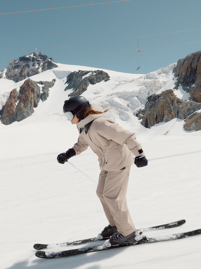 Model skiing downhill in a beige performance outfit on a sunlit slope, with rugged mountain cliffs and a clear blue sky in the background.