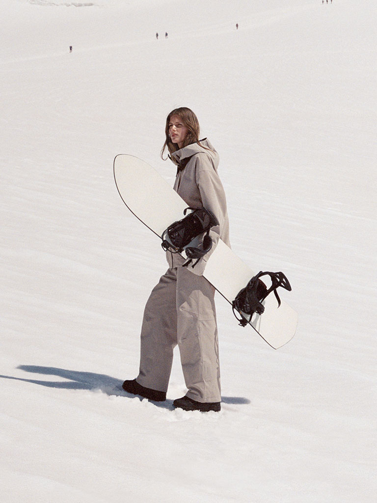 Model walking across a snowy slope holding a white snowboard, dressed in a light beige hooded snowsuit with black boots.